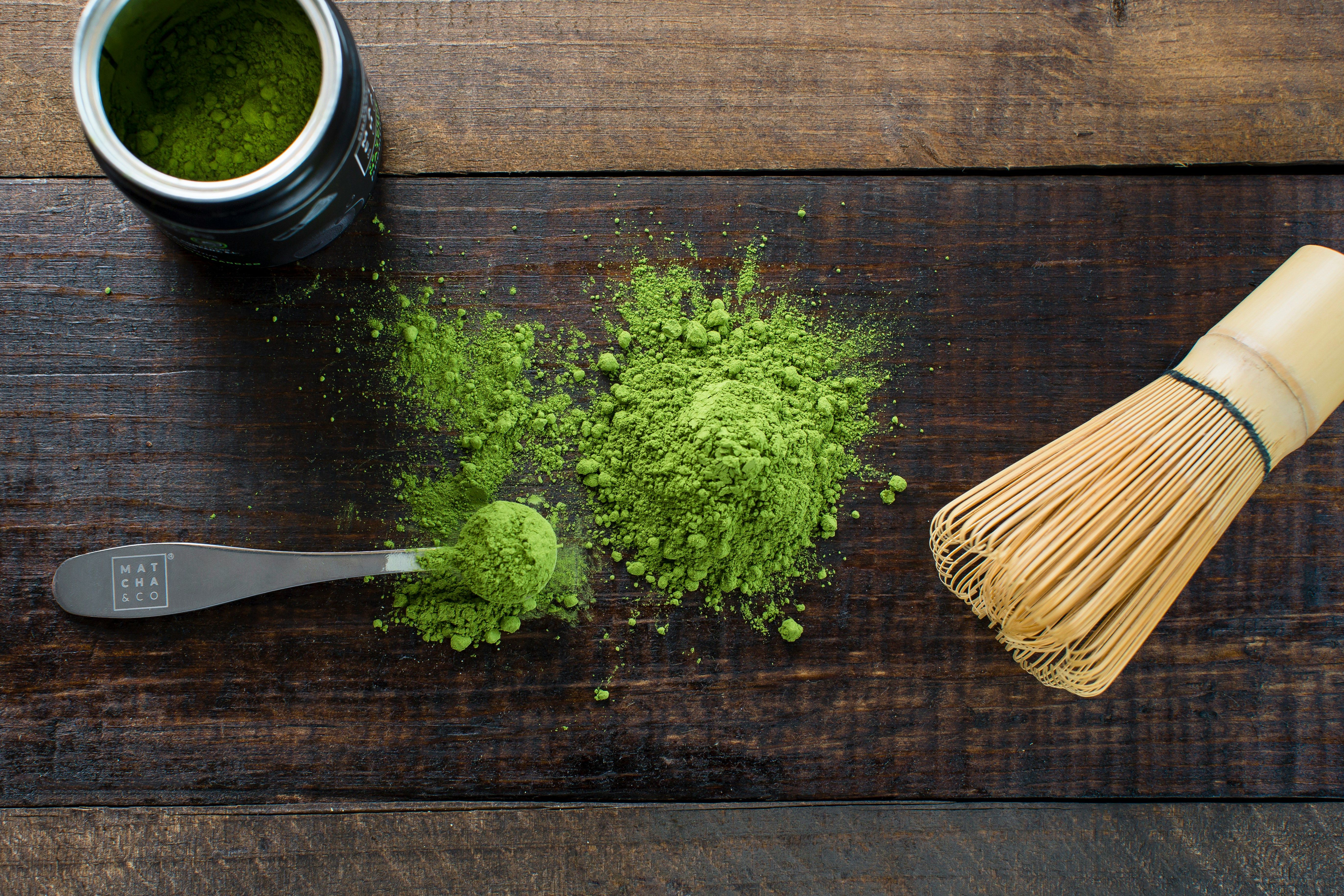 A spoonful of matcha place on a wooden tabletop with match sprinkled around the spoon.