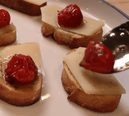 Step 3.1: Layer each toasted baguette slice with asiago cheese, tomato garlic confit, a slice of prosciutto, a sprig of basil, and drizzle with honey